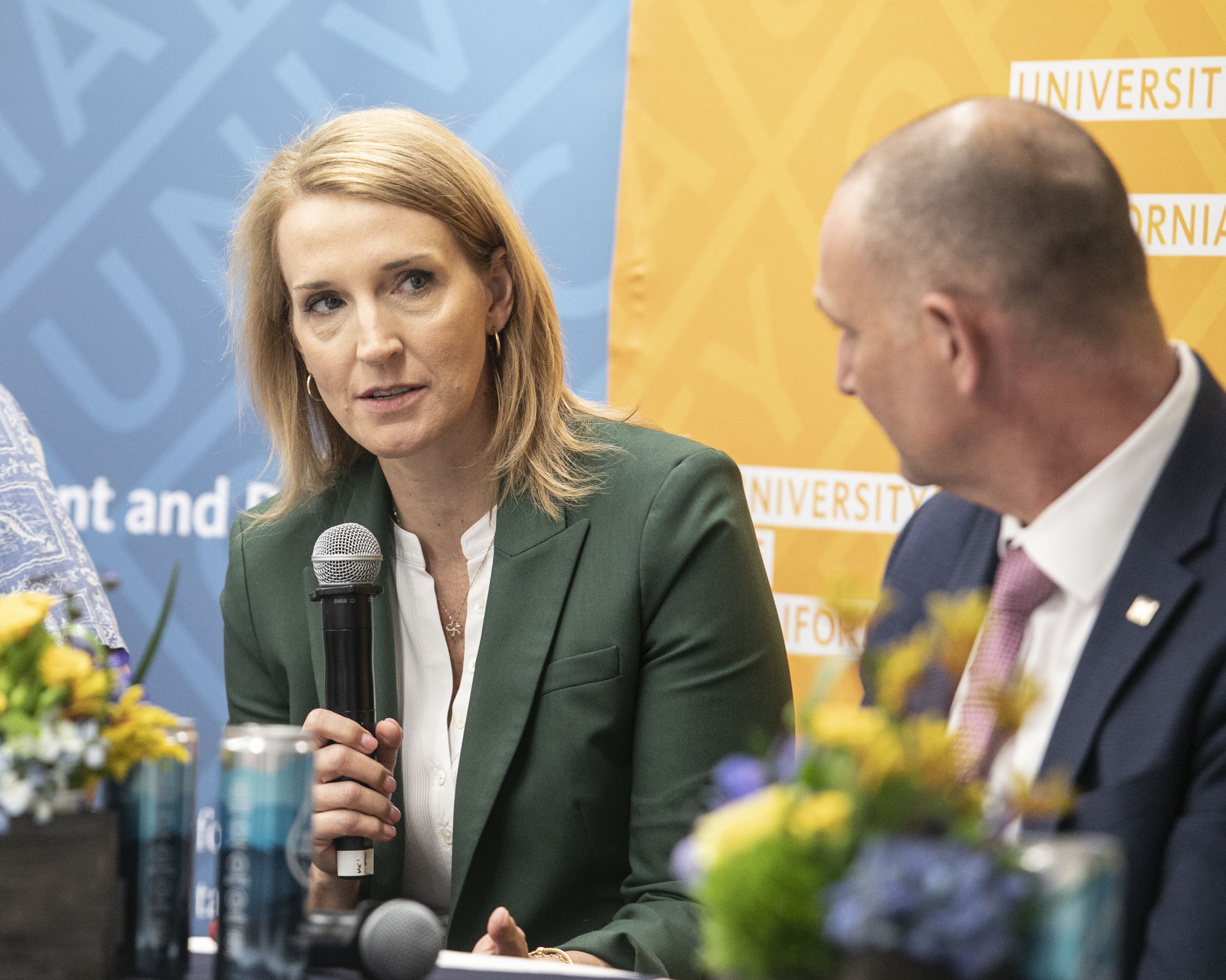 A woman in a green blazer speaks into a microphone during a panel discussion as another panelist listens, with University of California banners in the background.