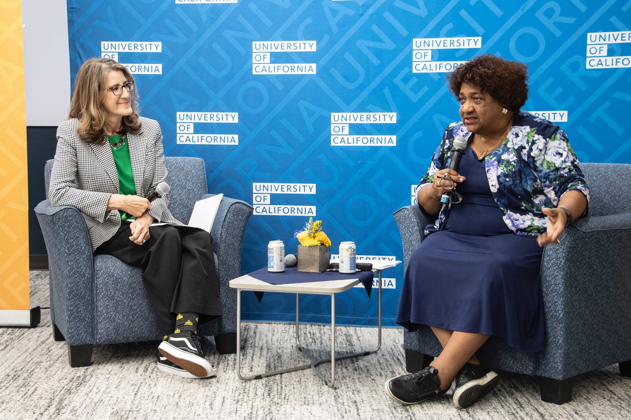 Two women sit in armchairs during a University of California speaker event. One holds a microphone and listens while the other speaks into a microphone, with a blue University of California backdrop behind them.